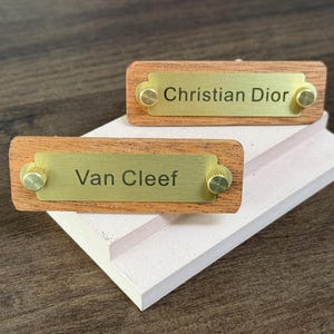 May include: Two wooden nameplate holders with brass accents. The top nameplate reads "Christian Dior" and the bottom nameplate reads "Van Cleef".