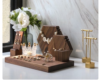 May include: A display of jewelry on wooden stands and a tray. The jewelry includes earrings, necklaces, rings, and a crown. The stands are various shapes and sizes, with a gold-colored earring stand. A bouquet of white flowers is in the background.
