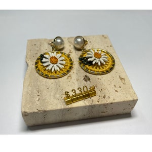 May include: A pair of gold earrings with white daisy flower accents and yellow thread detailing. The earrings are on a beige marble surface with a price tag that reads $330.00.