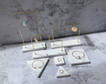 Può includere: Un set di cinque espositori in marmo bianco con cornici in metallo dorato. Gli espositori sono progettati per contenere gioielli e altri piccoli oggetti. Gli espositori sono disposti in uno schema sfalsato su uno sfondo grigio. Il testo "THE UNIQUE DISPLAY" è stampato nell'angolo in basso a destra dell'immagine.