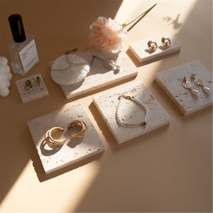 May include: Six small, square, light beige, textured, stone-like display trays with gold jewelry on them. The trays are arranged on a light beige surface.