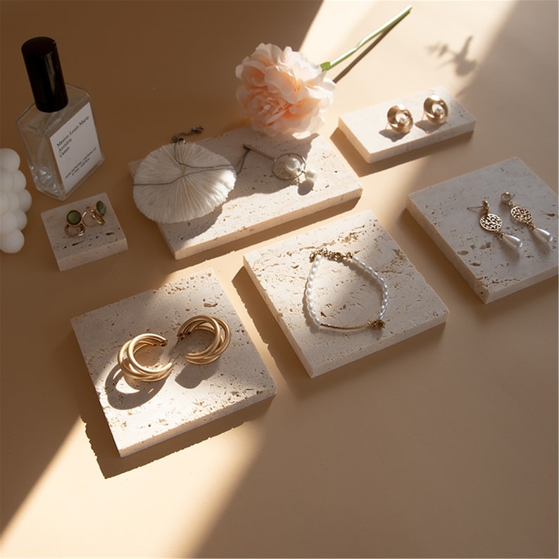 May include: Six small, square, light beige, textured, stone-like display trays with gold jewelry on them. The trays are arranged on a light beige surface.