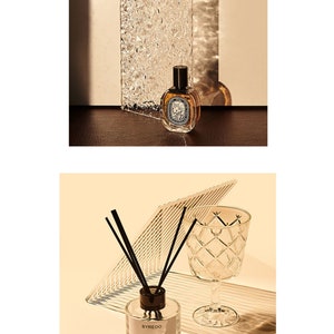 May include: Two glass bottles of Byredo Super Cedar perfume, one in a spray bottle and one in a diffuser bottle with black sticks. The bottles are on a brown surface with a clear glass object behind them.