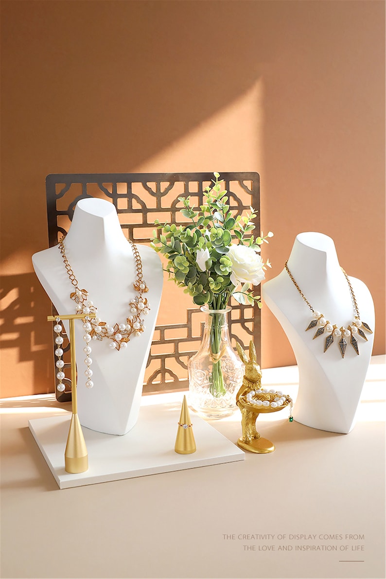 May include: A white jewellery display stand with two white mannequins wearing necklaces. The stand has a gold metal ring holder and a gold metal bunny figurine holding a small plate with a bracelet. The stand is on a white surface with a brown background.