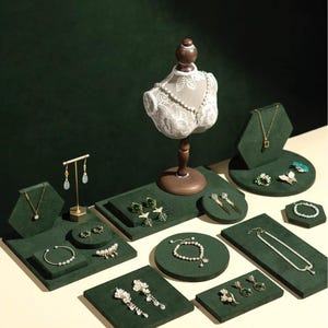 May include: A collection of jewellery displayed on green velvet platforms. The display includes necklaces, bracelets, earrings, and rings. A mannequin bust with a pearl necklace is central. The jewellery is set against a dark green backdrop.