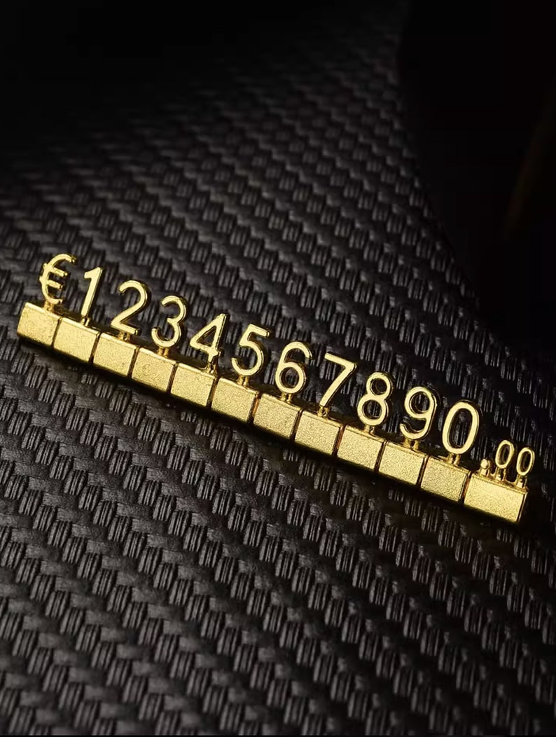 May include: Gold-colored number tiles with the euro symbol and numbers 1 through 0. The tiles are arranged in a row on a black textured surface.