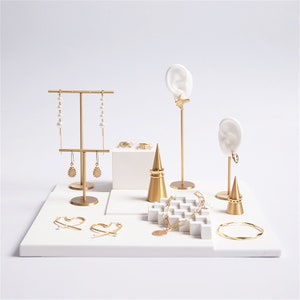 May include: Gold jewelry display stand with white base. The stand features a variety of earring holders, a ring cone, and a necklace holder. The stand is perfect for showcasing jewelry.