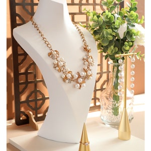 May include: A white jewellery display stand with a gold necklace, earrings, and ring. The necklace features a gold chain with white pearls and gold leaf accents. The earrings are long and dangle with white pearls. The ring is on a gold cone-shaped stand.