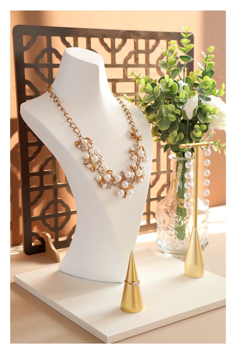May include: A white jewellery display stand with a gold necklace, earrings, and ring. The necklace features a gold chain with white pearls and gold leaf accents. The earrings are long and dangle with white pearls. The ring is on a gold cone-shaped stand.