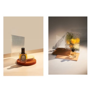 May include: A brown glass bottle of perfume with a black cap sits on a brown and beige circular platform. The bottle is in front of a textured glass panel.