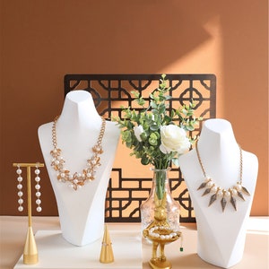 May include: Two white jewellery display busts with necklaces, a gold ring holder, a gold earring stand with pearl earrings, and a gold decorative stand with a pearl bracelet. The stands are on a white surface with a brown background.