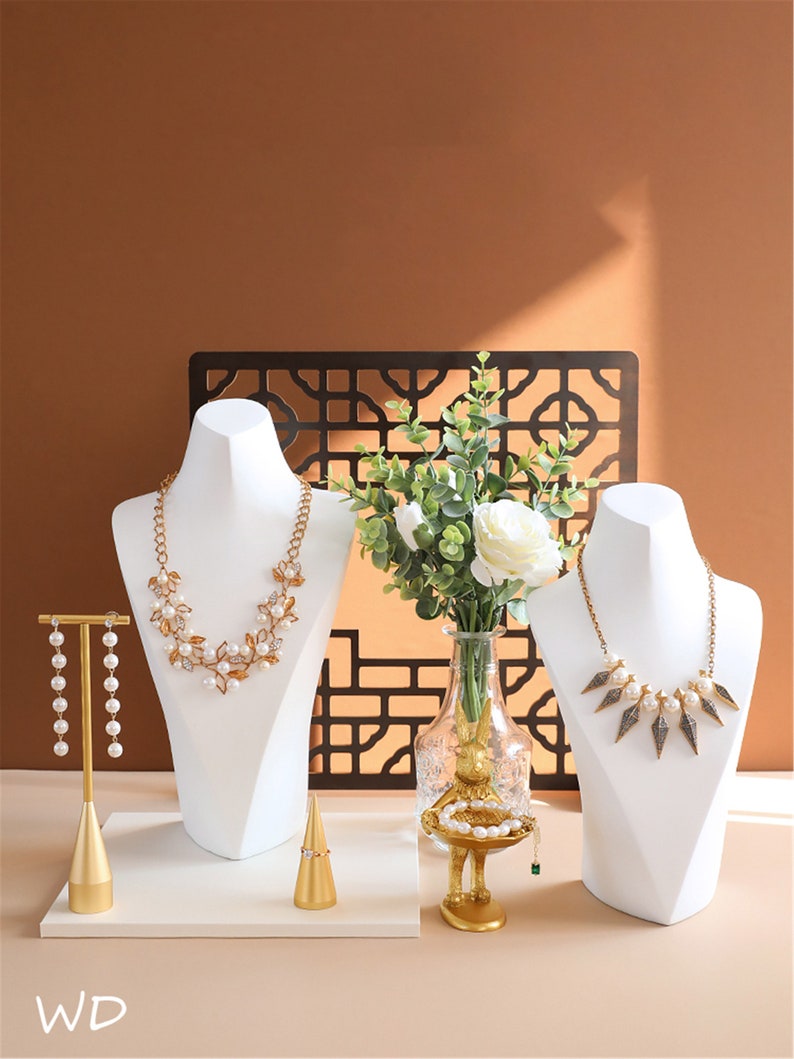 May include: Two white jewellery display busts with necklaces, a gold ring holder, a gold earring stand with pearl earrings, and a gold decorative stand with a pearl bracelet. The stands are on a white surface with a brown background.