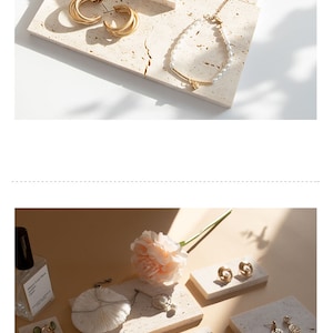 May include: A set of jewelry display stands made of beige travertine stone. The stands are arranged in a staggered pattern and feature various gold and pearl jewelry pieces, including earrings, necklaces, and rings. A pink flower is placed on top of the stands.