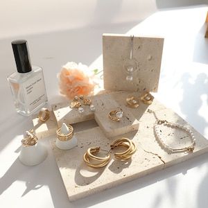 May include: A collection of gold jewelry displayed on small, beige, textured stone squares. The jewelry includes earrings, rings, a necklace, and a bracelet. The jewelry features pearls and gold accents.