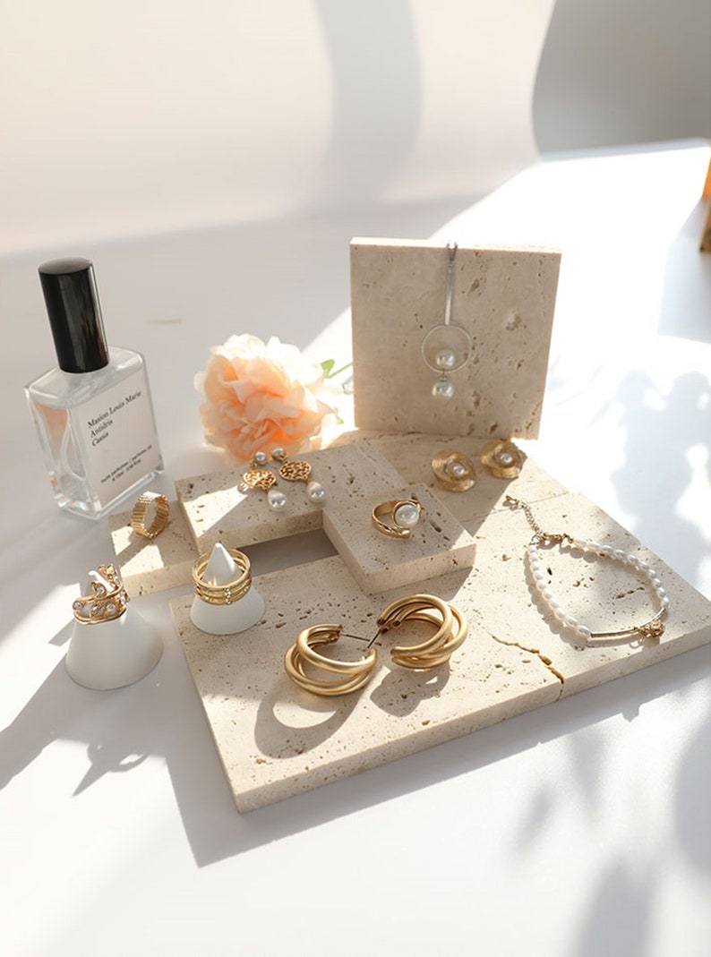 May include: A collection of gold jewelry displayed on small, beige, textured stone squares. The jewelry includes earrings, rings, a necklace, and a bracelet. The jewelry features pearls and gold accents.