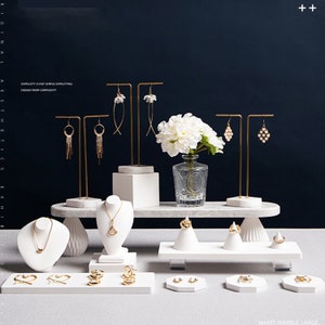 May include: White marble jewelry display stand with gold metal earring holders and necklace stands. The display stand has a tiered design with a rectangular base and two smaller tiers. The stand is decorated with a white flower arrangement in a clear glass vase.