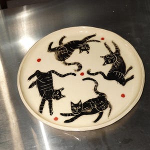 Może przedstawiać: Okrągły talerz ceramiczny z kremowym szkliwem, ozdobiony czterema ilustracjami czarnych kotów w różnych zabawnych pozach. Koty mają wygrawerowane detale liniowe, a pomiędzy nimi na powierzchni znajdują się małe czerwone kropki.