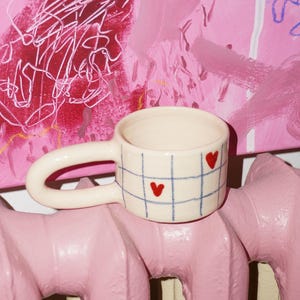 Puede incluir: Una taza de cerámica de color crema con un patrón de cuadrícula azul y pequeños iconos de corazones rojos. La taza tiene un asa ancha y redondeada que se extiende desde el lateral y un cuerpo cilíndrico. El diseño es simple y geométrico.