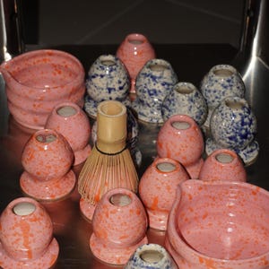Può includere: Una collezione di ciotole per matcha e supporti per fruste in ceramica con smalto maculato rosa e arancione o blu e bianco, accompagnata da una singola frusta per matcha in bambù marrone chiaro con setole sottili e flessibili.