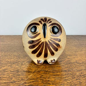 May include: A round ceramic owl figurine featuring a speckled beige body with dark brown painted feather patterns. The owl has two large, glossy black circular eyes and a prominent, vertical black beak centred on its face, with two small feet.