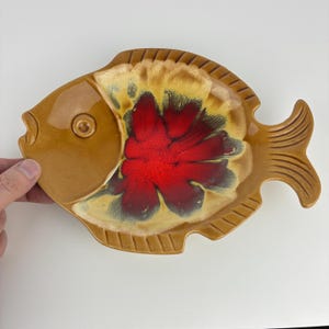 Peut inclure: Un plat en céramique en forme de poisson avec un extérieur marron et un design intérieur rouge et jaune vif. Le plat a une surface texturée et est conçu avec une queue et des nageoires. L'objet est décoratif.