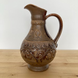 May include: A brown ceramic pitcher with a handle and spout. The pitcher has embossed grape and leaf designs, and the word "VINO" is embossed on the front. The pitcher is on a wooden surface.