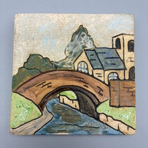 May include: Square ceramic tile featuring a hand-painted landscape scene. The artwork depicts a brown bridge arching over a blue river, with buildings and green foliage in the background. The tile has a textured finish.