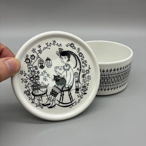 Könnte beinhalten: Ein weißes Keramikschalen- und Deckelset. Der Deckel zeigt eine Schwarz-Weiß-Illustration einer Person, die mit einer Katze sitzt, umgeben von floralen und dekorativen Elementen. Die Schüssel hat ein schwarzes geometrisches Muster.