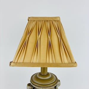 May include: A gold-coloured lampshade with a pleated design. The shade is trapezoidal and sits atop a decorative lamp base with a gold and white finish. The lamp is set against a white background.