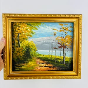 May include: Oil painting of a landscape scene with a path leading to a body of water. The artwork features trees with yellow and orange foliage, a blue sky, and a gold-colored ornate frame.