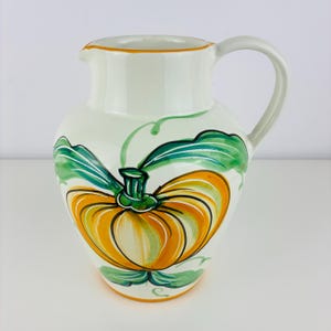 May include: White ceramic pitcher with a large orange and green pumpkin design. The pitcher has a curved handle and a spout. Green leaves and vines surround the pumpkin. The rim is trimmed with orange.
