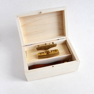 Puede incluir: Una caja de madera abierta que revela una herramienta de latón con un mango de madera. La herramienta tiene un borde curvo y dentado, probablemente para trabajos en cuero. La caja es de madera clara y tiene una tapa con bisagras.