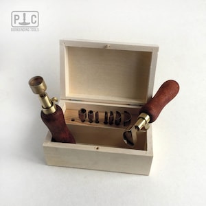 May include: A set of bookbinding tools in a wooden box. The tools include a variety of punches and a leather stitching awl. The tools are made of brass and have wooden handles.