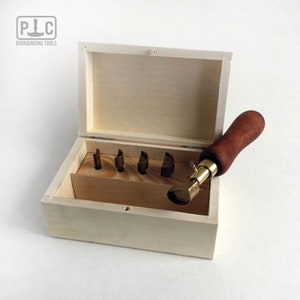 Könnte beinhalten: Ein Satz Buchbinderwerkzeuge in einer Holzkiste. Die Werkzeuge umfassen einen Lederglätter mit einem Holzgriff und einen Satz Messingstanzwerkzeuge.