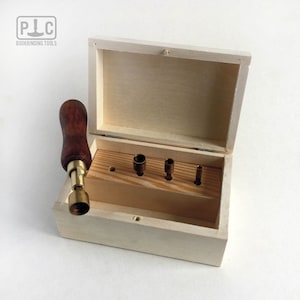 Peut inclure: Un ensemble d'outils de reliure dans une boîte en bois. Les outils comprennent un poinçon avec un manche en bois et cinq poinçons de différentes tailles. La boîte est en bois clair et possède un couvercle à charnières.