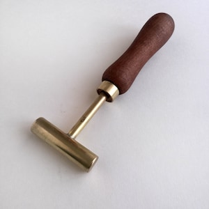 Puede incluir: Una herramienta en forma de T de latón con un mango de madera. La herramienta tiene una pequeña inscripción que dice "1843".