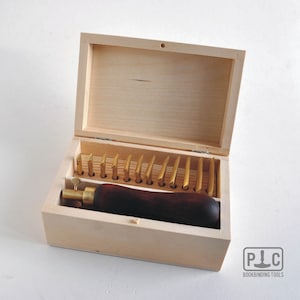 Może przedstawiać: Drewniane pudełko z zestawem 16 złotych narzędzi do introligatorstwa. Narzędzia są ułożone w rzędach i są utrzymywane na miejscu przez drewnianą ramę. Pudełko jest otwarte, a narzędzia są widoczne. Pudełko jest oznaczone "P.I.C. Bookbinding Tools".