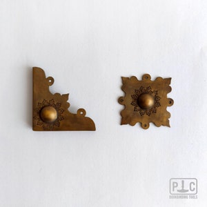 Può includere: Due pezzi di ferramenta antica per rilegatura in ottone. Uno è un angolo, l'altro è una piastra quadrata. Entrambi hanno un disegno centrale in rilievo e fori di montaggio. L'angolo misura circa 5 cm di lunghezza.