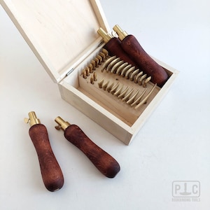 May include: A set of bookbinding tools in a wooden box. The tools are made of brass and have wooden handles. The box is open and the tools are visible. The tools are used for scoring and cutting leather.
