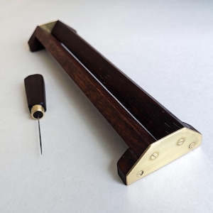 Może przedstawiać: Ciemne drewniane narzędzie do wyrobu galanterii skórzanej z mosiężnymi akcentami. Narzędzie ma długą, prostokątną ramę z kanałem i małe szydło z ciemnym uchwytem. Rama ma około 30 cm długości.