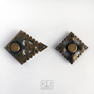 Könnte beinhalten: Zwei verzierte Messing-Buchbindeecken mit aufwendigen floralen Mustern und einem runden Mittelstück. Die Ecken sind rautenförmig und haben einen gewellten Rand.