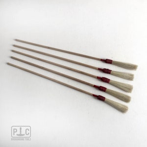 Könnte beinhalten: Sechs Buchbindepinsel mit Naturborsten und Holzstielen. Die Stiele sind hellbraun und die Borsten hellbeige. Die Basis der Borsten ist mit einem dunkelroten Material umwickelt. Das Logo "PLC BOOKBINDING TOOLS" befindet sich unten links.