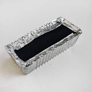 Puede incluir: Un recipiente rectangular de papel de aluminio con un objeto negro en su interior. El papel de aluminio está arrugado en los bordes. El recipiente está sobre una superficie blanca. El objeto negro parece ser un bloque sólido.