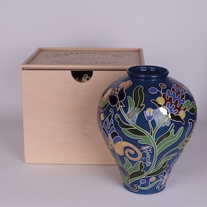 May include: A blue ceramic vase with floral designs in green, yellow, and purple. The vase has a round shape and a narrow neck. A wooden box is in the background. The vase has the word "Helmansla" written on it.