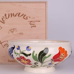 Pode incluir: Uma tigela de cerâmica creme com um desenho floral em vermelho, laranja, azul e verde. A tigela tem uma borda larga e uma base de pedestal. Uma caixa de madeira com texto está no fundo.