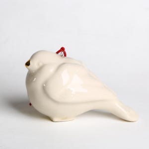 Puede incluir: Adorno de cerámica con forma de paloma de color blanco crema. El ave tiene un pico dorado y una cuerda roja para colgar. La paloma es un símbolo de paz y se utiliza a menudo como objeto decorativo.