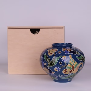 Peut inclure: Un vase en céramique bleu foncé avec un motif floral dans les tons de vert, jaune, marron et violet. Le vase a une forme arrondie et une ouverture étroite. Une boîte en bois est en arrière-plan.