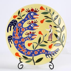 May include: A decorative ceramic plate with a yellow background. The plate features a blue serpent-like creature with orange accents, a small figure, and floral elements. The plate is displayed on a black metal stand.