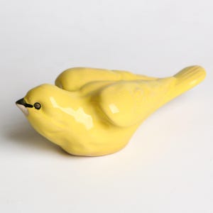 以下が含まれることがあります： 光沢のある仕上がりのセラミック製の黄色い鳥の置物。鳥は黒い嘴と目の詳細を持っています。翼はわずかに持ち上げられ、尾は広がっています。置物は白い背景に設定されています。