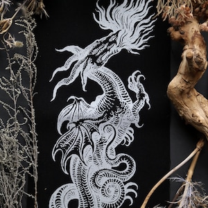 Puede incluir: Impresión en lienzo negro con una detallada ilustración en blanco de un dragón con alas, cuerpo serpentino y melena fluida. La obra está enmarcada por plantas y ramas secas, creando una estética gótica.